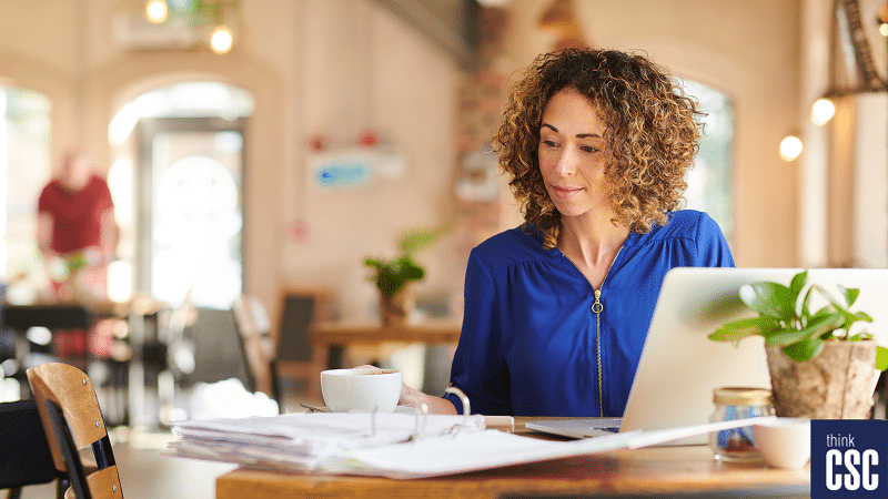 woman in blue shirt with curly hair sitting in cafe working with laptop open and a cup of coffee